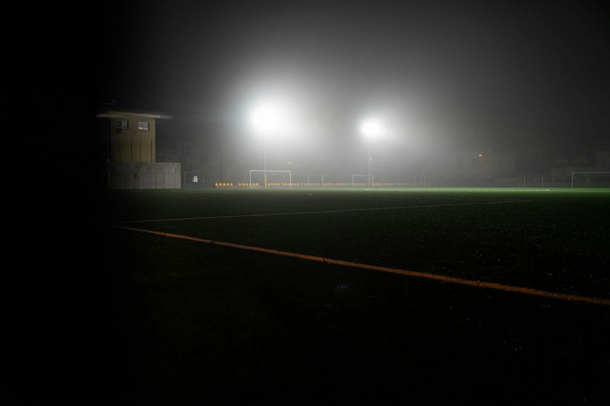 An empty field at night in fog, two floodlights glowing through the haze and a chalk line running across the grass.
