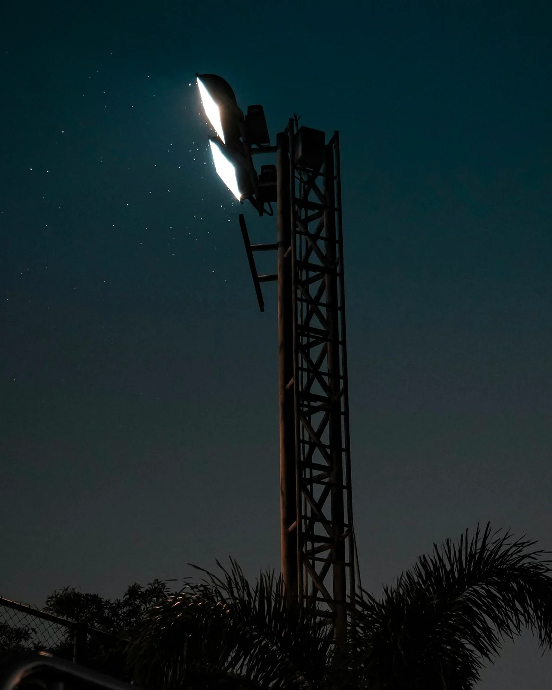 A floodlight tower lit against a dark evening sky, with insects caught in its beam.