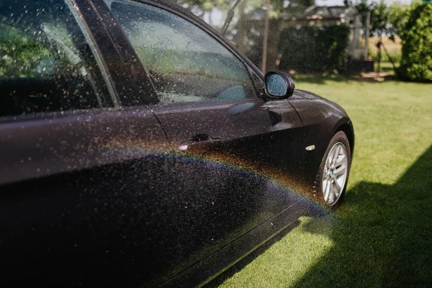 A small rainbow in the spray as a dark car is rinsed on a lawn
