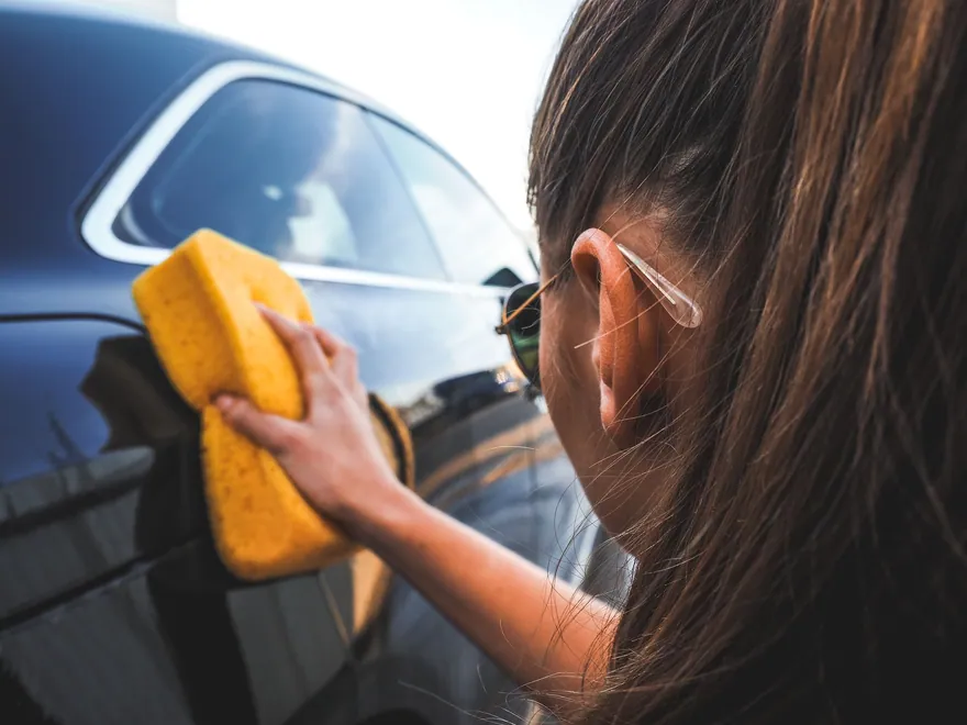 A woman washing the side of a dark car with a yellow sponge