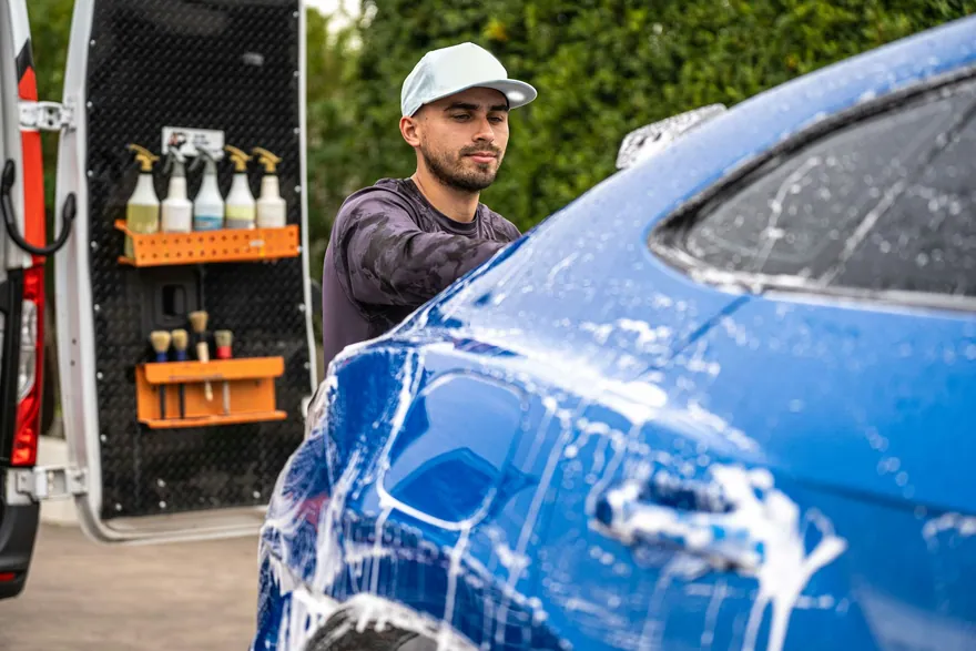 A mobile detailer washing a blue car beside his van, with spray bottles and brushes racked on the door