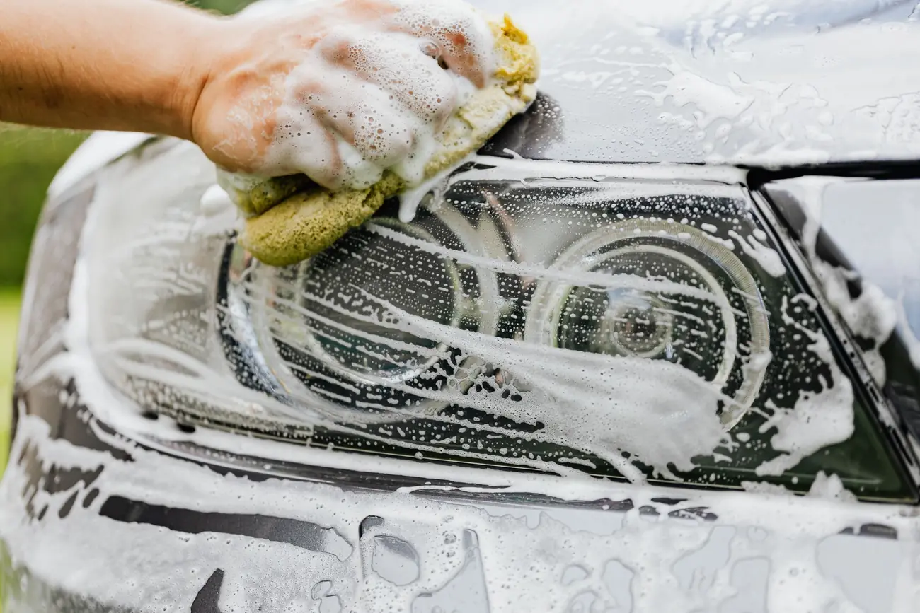 A hand washing a car headlight with a soapy yellow sponge