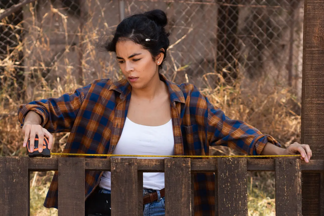 A woman in a plaid shirt measuring the top of a wooden fence with a tape measure
