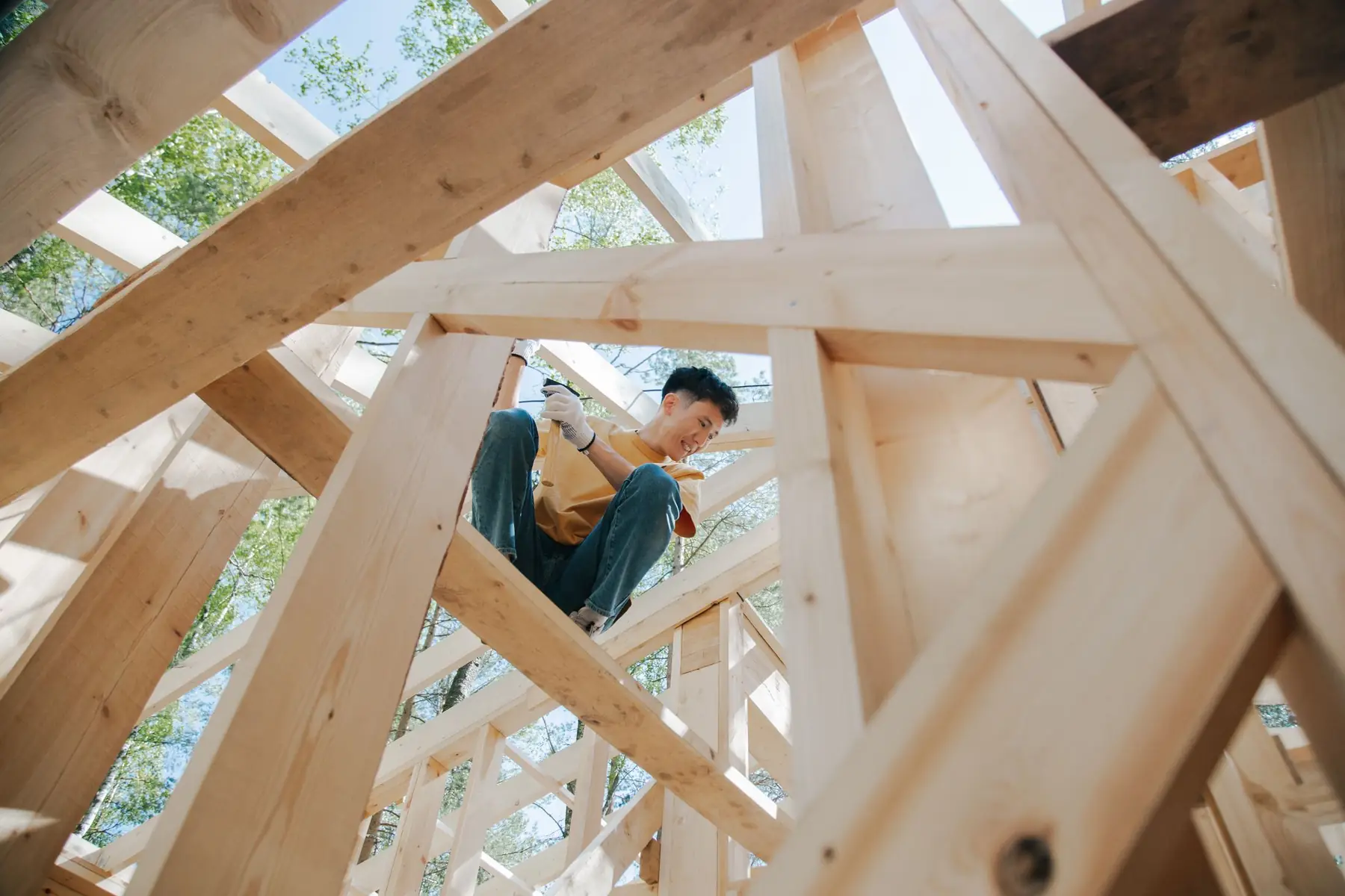 Looking up through the timber frame of a house under construction, a carpenter sitting on a beam