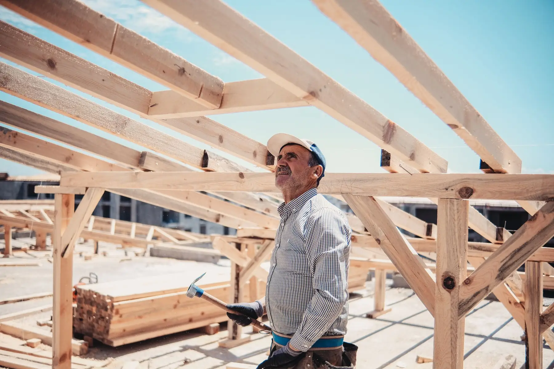 A builder in a cap and work shirt standing under new timber rafters, hammer in hand, looking up at the frame