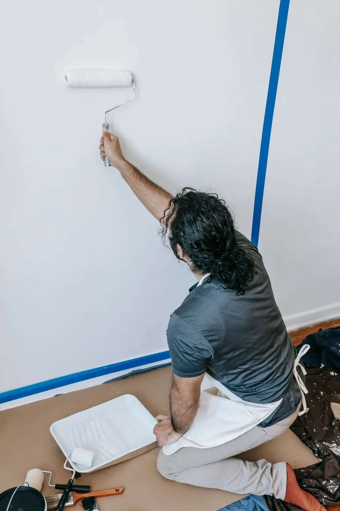A painter on one knee rolling white paint up a wall beside blue masking tape