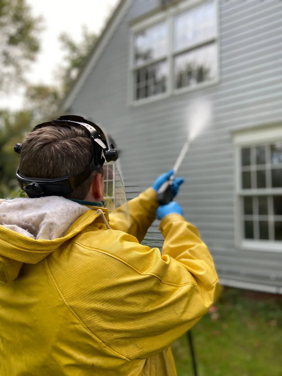 A worker in a yellow rain jacket pressure washing gray house siding