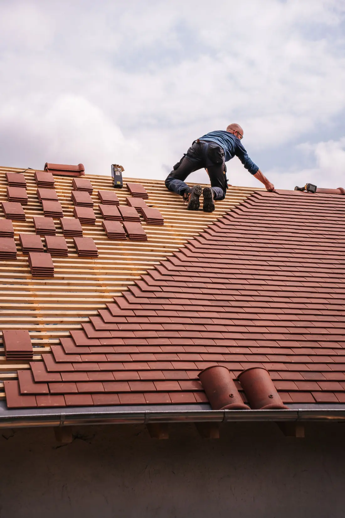 A roofer kneeling on a roof, laying red clay tiles over wooden battens