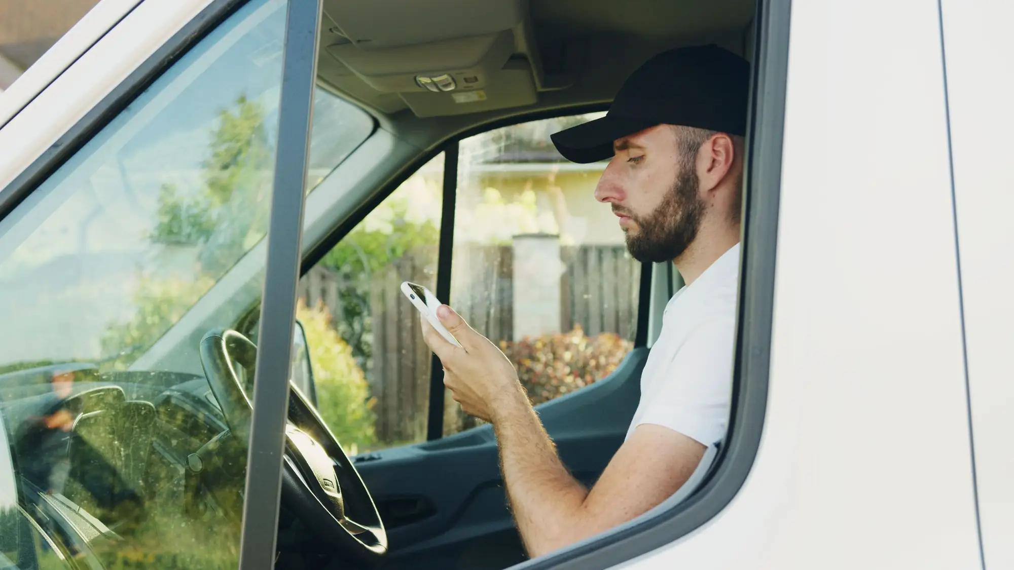 A contractor in a cap sitting in the driver's seat of a white work van, looking at his phone