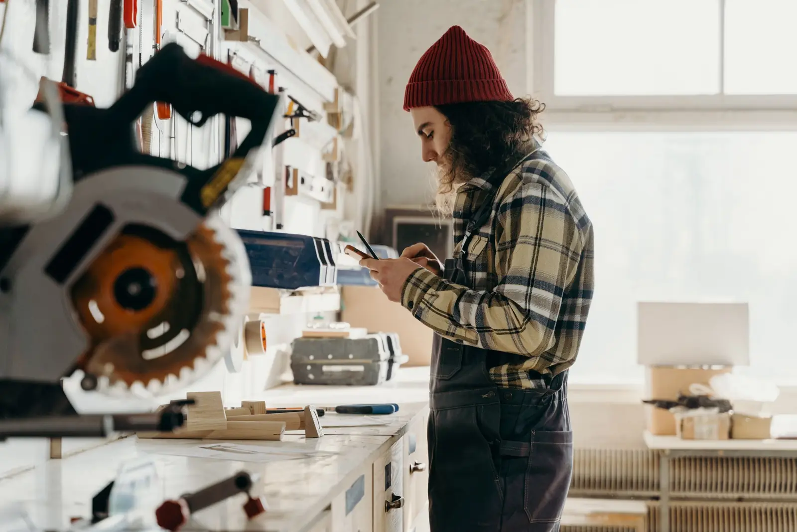 A woodworker in a plaid shirt and red beanie looking at a phone beside a miter saw in a bright workshop