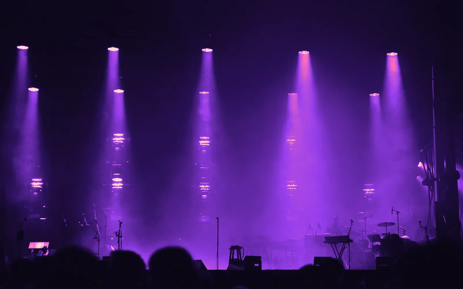 Beams of stage light through haze above an empty stage, seen over the heads of a waiting crowd