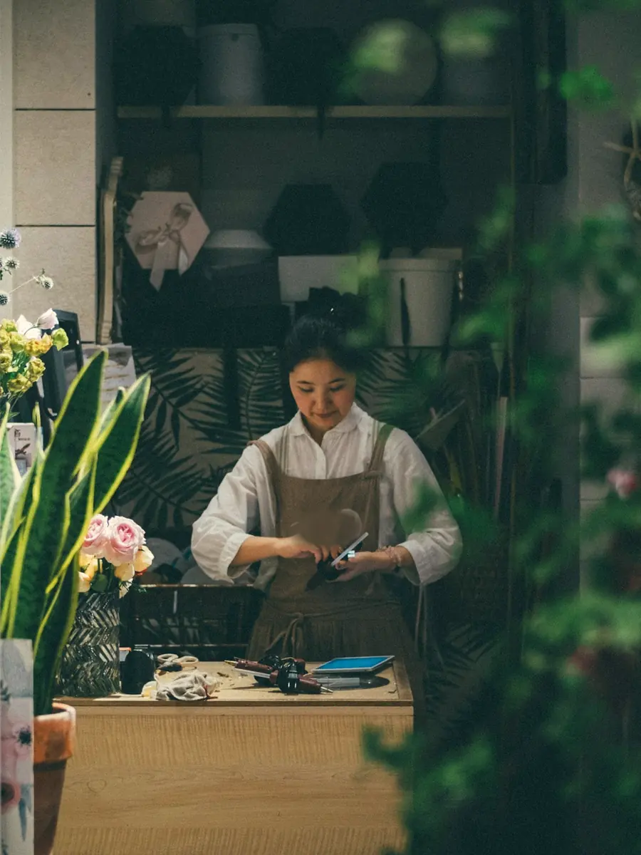 A florist in an apron working at her counter, seen through leaves and flowers
