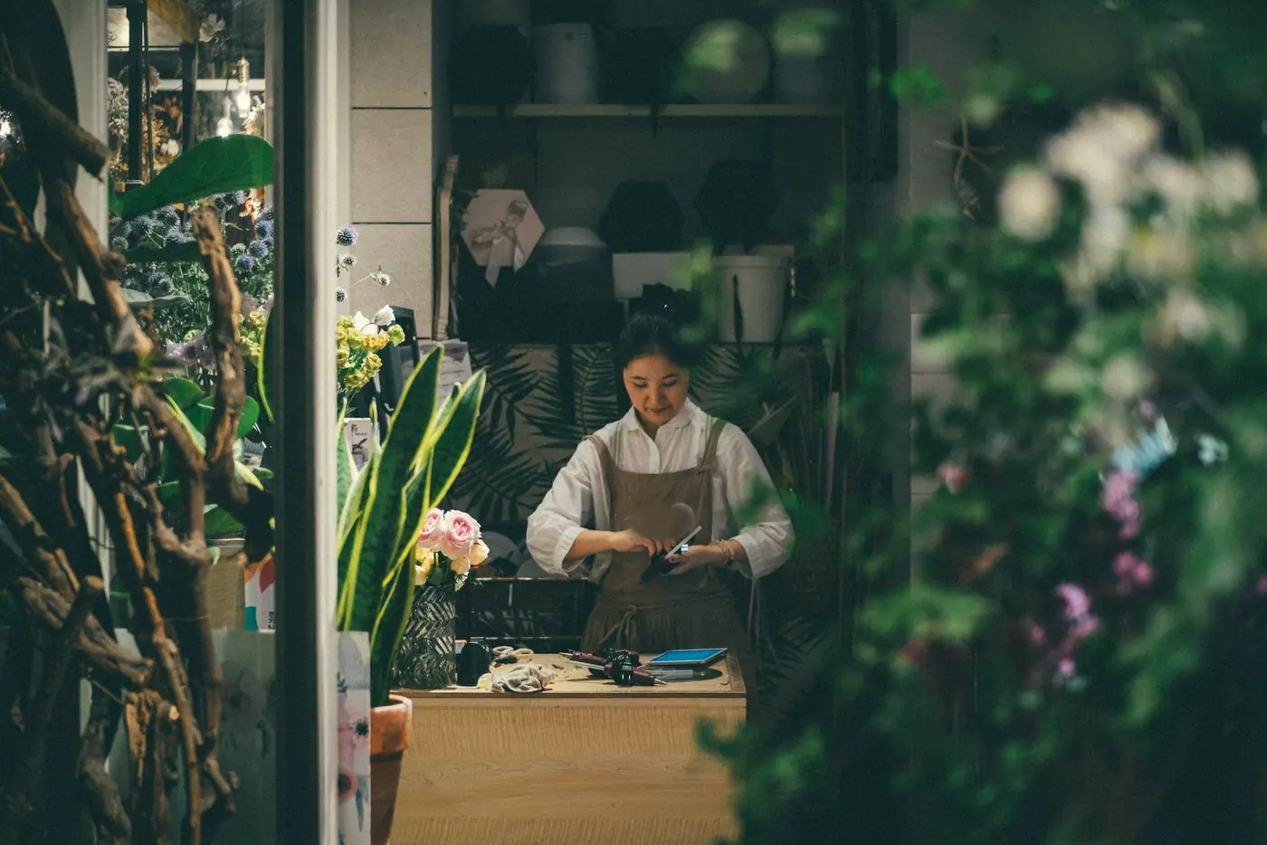 A florist in an apron working at her counter, seen through leaves and flowers