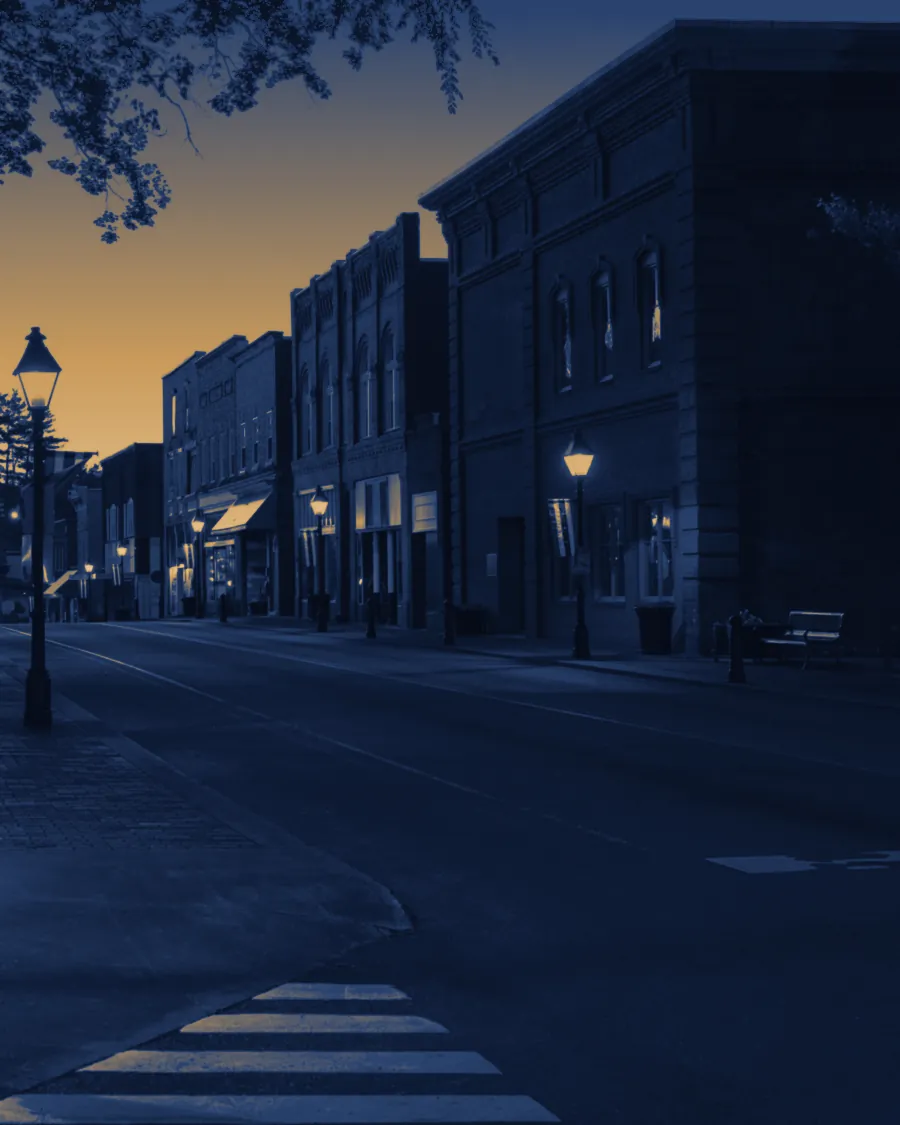 A small-town main street at dusk: brick shop fronts, street lamps coming on, an empty road.